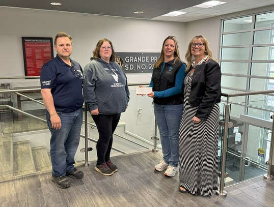 Four people stand together inside the Grande Prairie Public School Division office, smiling at the camera. From left to right: a man in a navy tournament t-shirt, a woman in a grey Iceberg hoodie, a woman in a black vest handing over a donation cheque, an