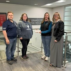 Four people stand together inside the Grande Prairie Public School Division office, smiling at the camera. From left to right: a man in a navy tournament t-shirt, a woman in a grey Iceberg hoodie, a woman in a black vest handing over a donation cheque, an