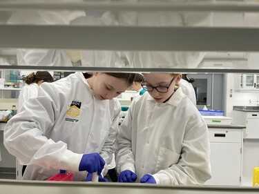 2 students in white science jumpsuits and purple gloves on doing a science experiment